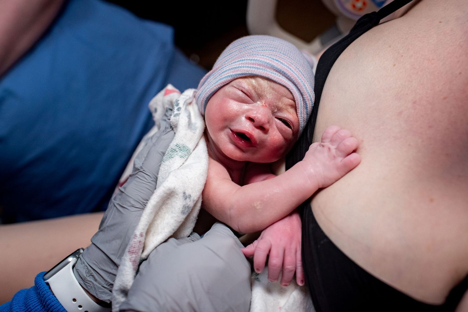 Newborn baby in gray hat crying on mother's chest moments after birth in hospital.