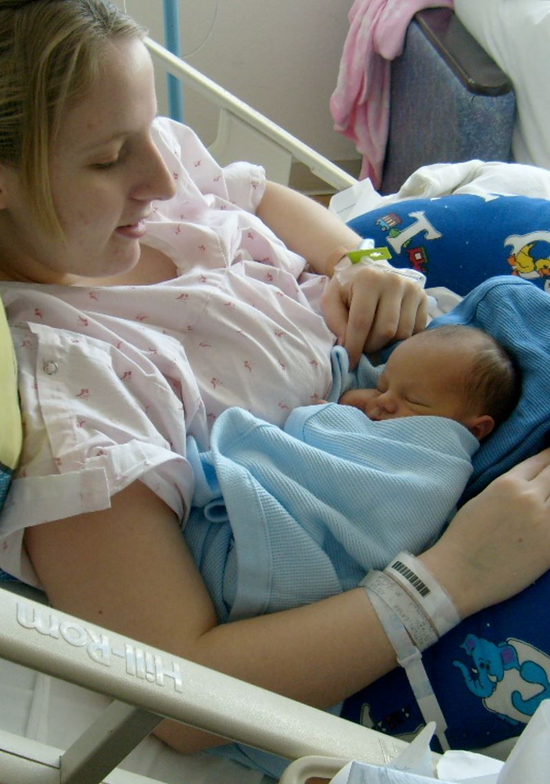 New mother in hospital gown cradling sleeping newborn baby wrapped in blue blanket in maternity ward.