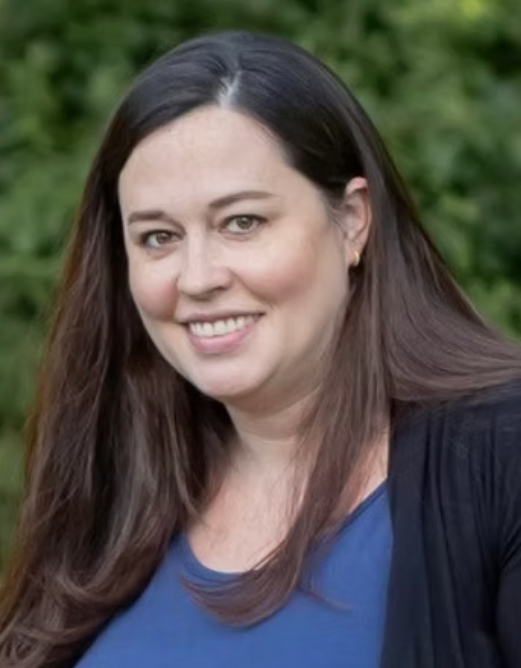 Smiling woman with long dark hair wearing a blue top and black cardigan outdoors with green foliage background.