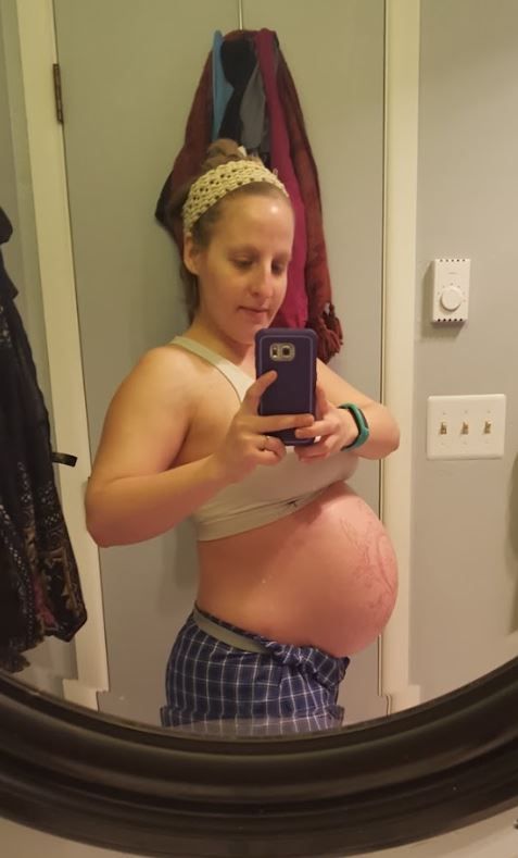 Pregnant woman taking mirror selfie showing large baby bump in bathroom wearing sports bra and plaid shorts.