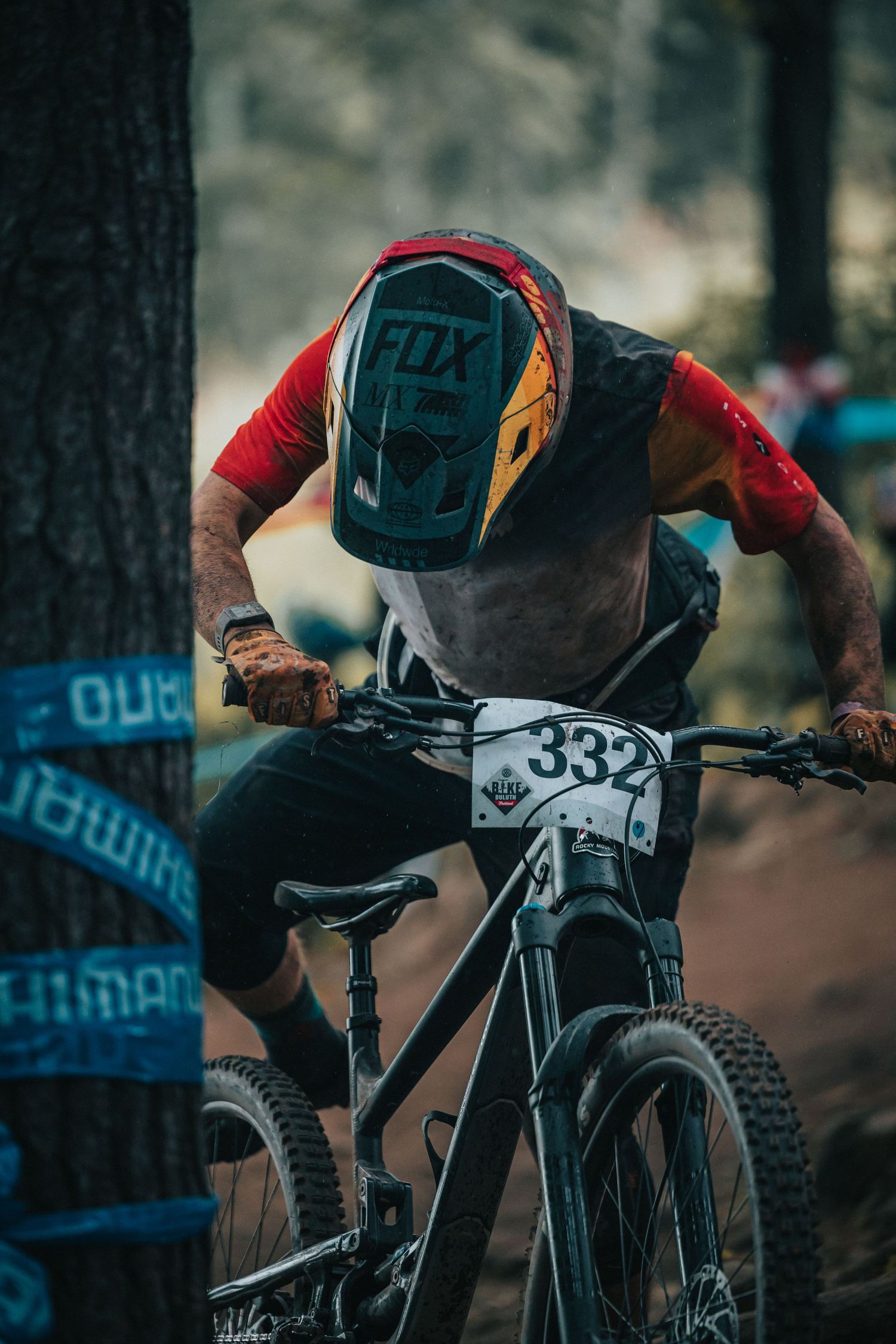 Racer #332 navigates a muddy forest trail on a mountain bike wearing a Fox helmet during a downhill race.