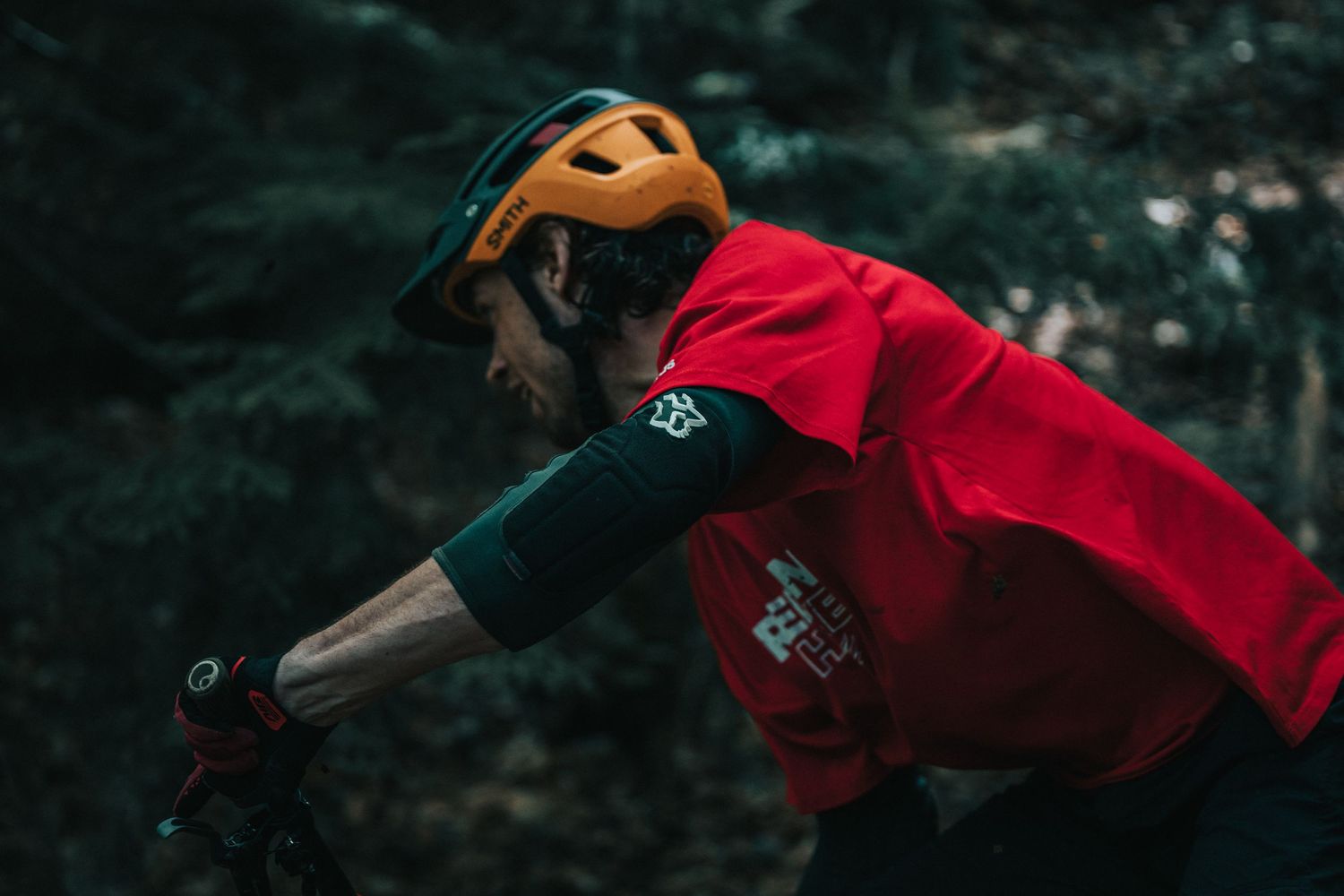Male mountain biker in red jersey and orange helmet resting on bike in forest.