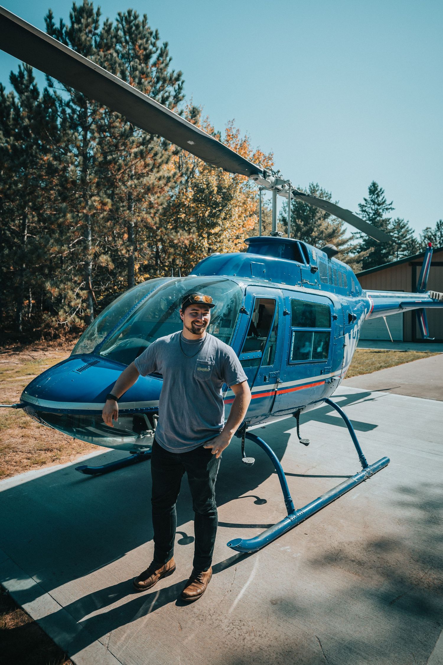 Smiling man in grey t-shirt poses in front of a blue Bell 206 helicopter on a sunny day.