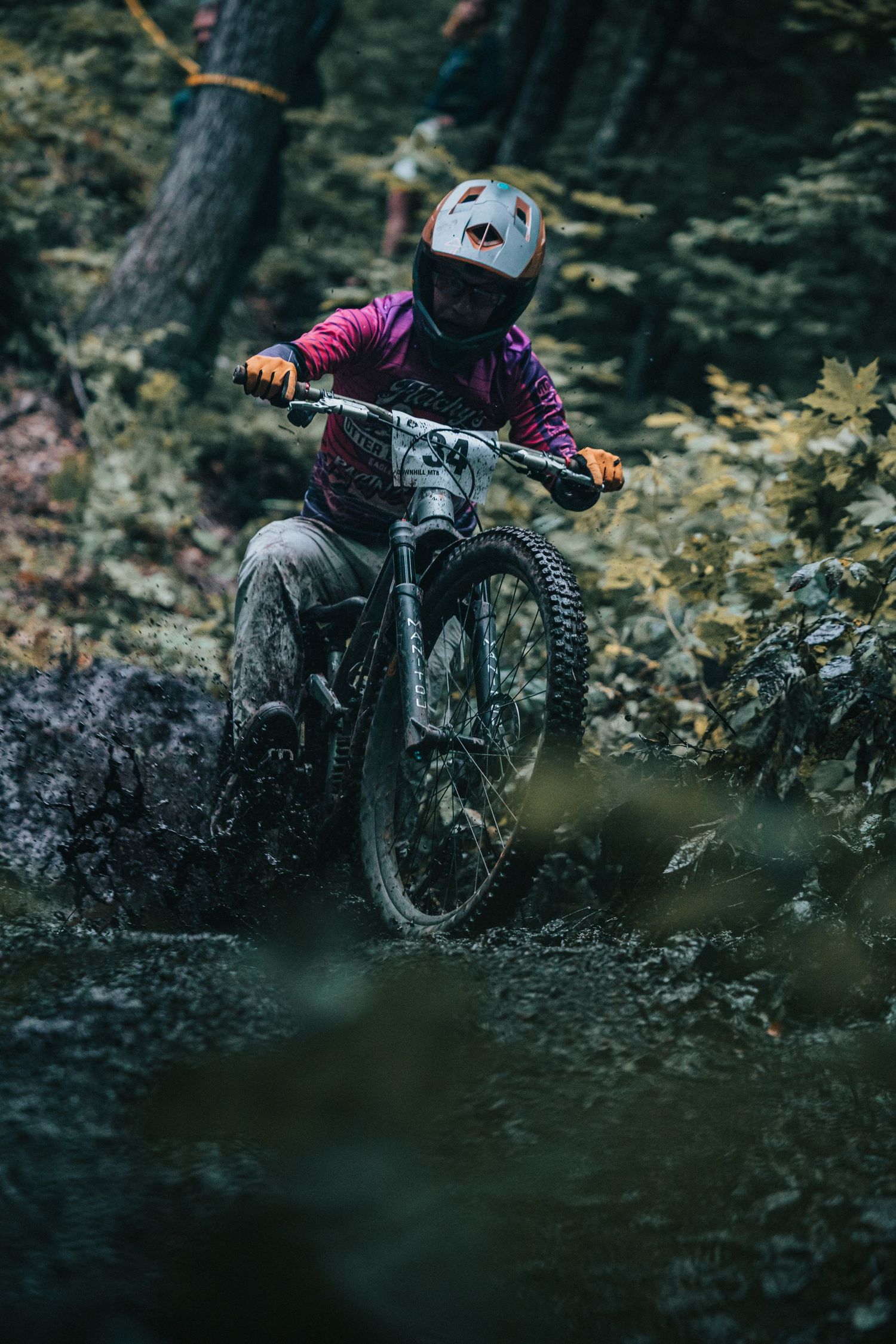Mountain biker in purple jersey powers through muddy forest trail on a full-suspension bike in a race.