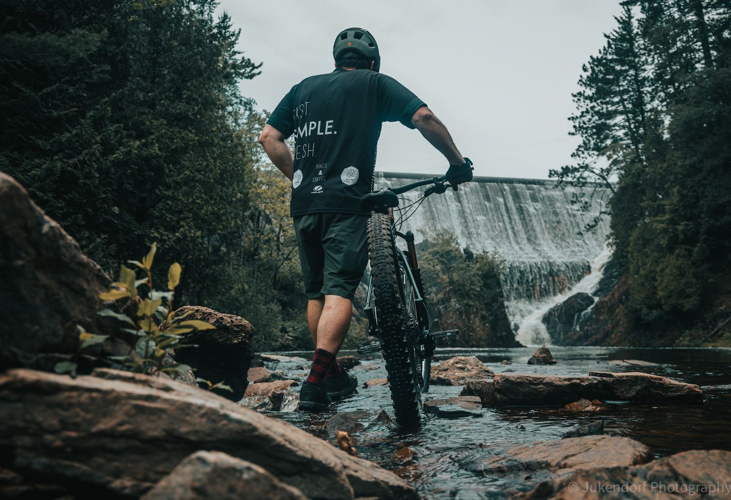 Mountain biker standing in rocky stream with waterfall and dense forest in background.