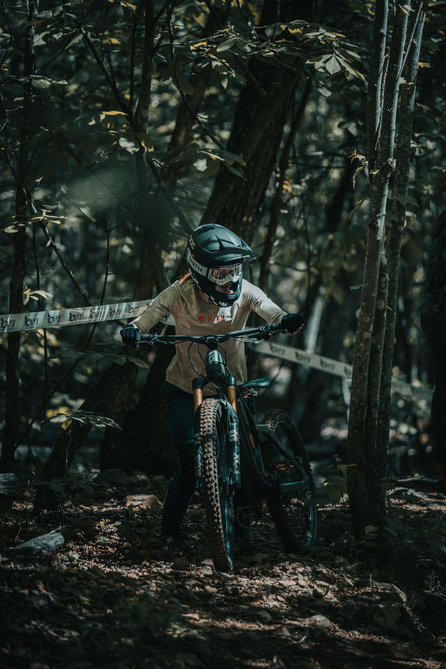 Mountain biker in protective gear rides through a dark wooded trail marked with tape.