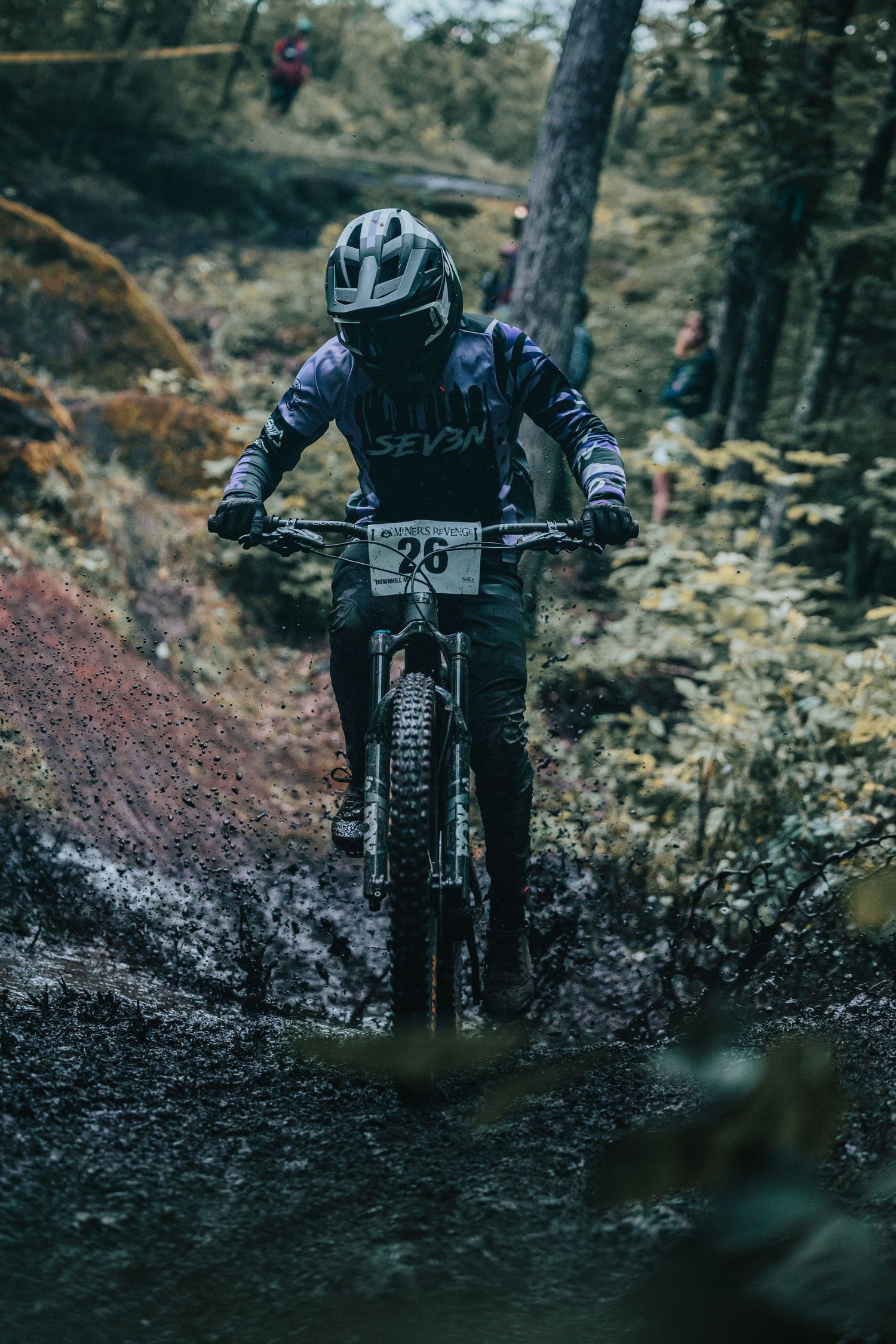 Mountain biker #26 in Seven gear descends a muddy forest trail during a downhill enduro race.