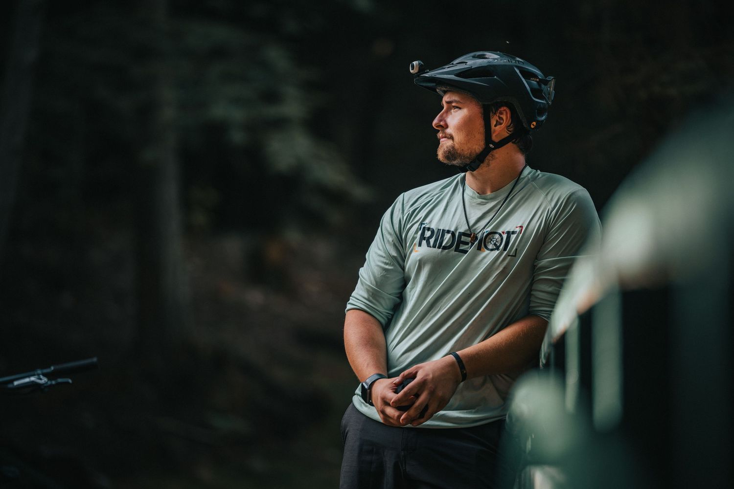 Mountain biker in gray RIDE shirt and helmet stands thoughtfully in a dark forest setting.
