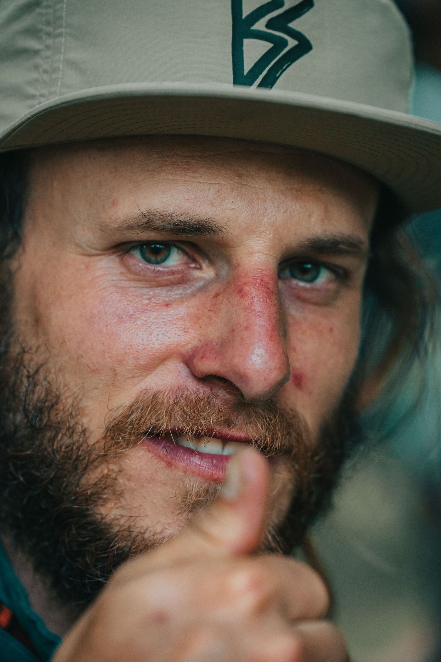 Close-up portrait of bearded man with blue eyes wearing tan cap pointing finger forward.