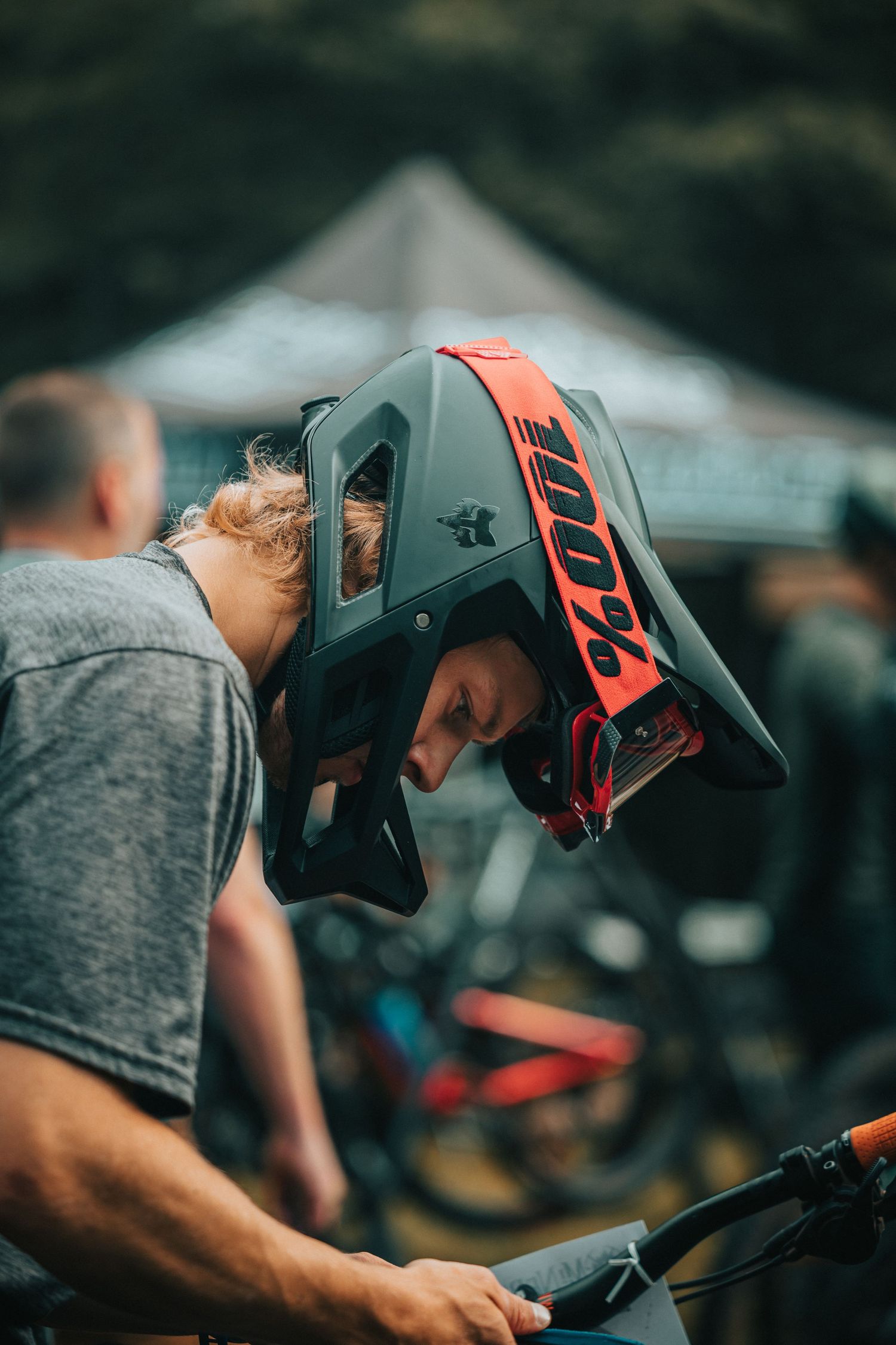 Young mountain biker wearing a red and grey 100% full-face helmet adjusts bike at an outdoor cycling event.