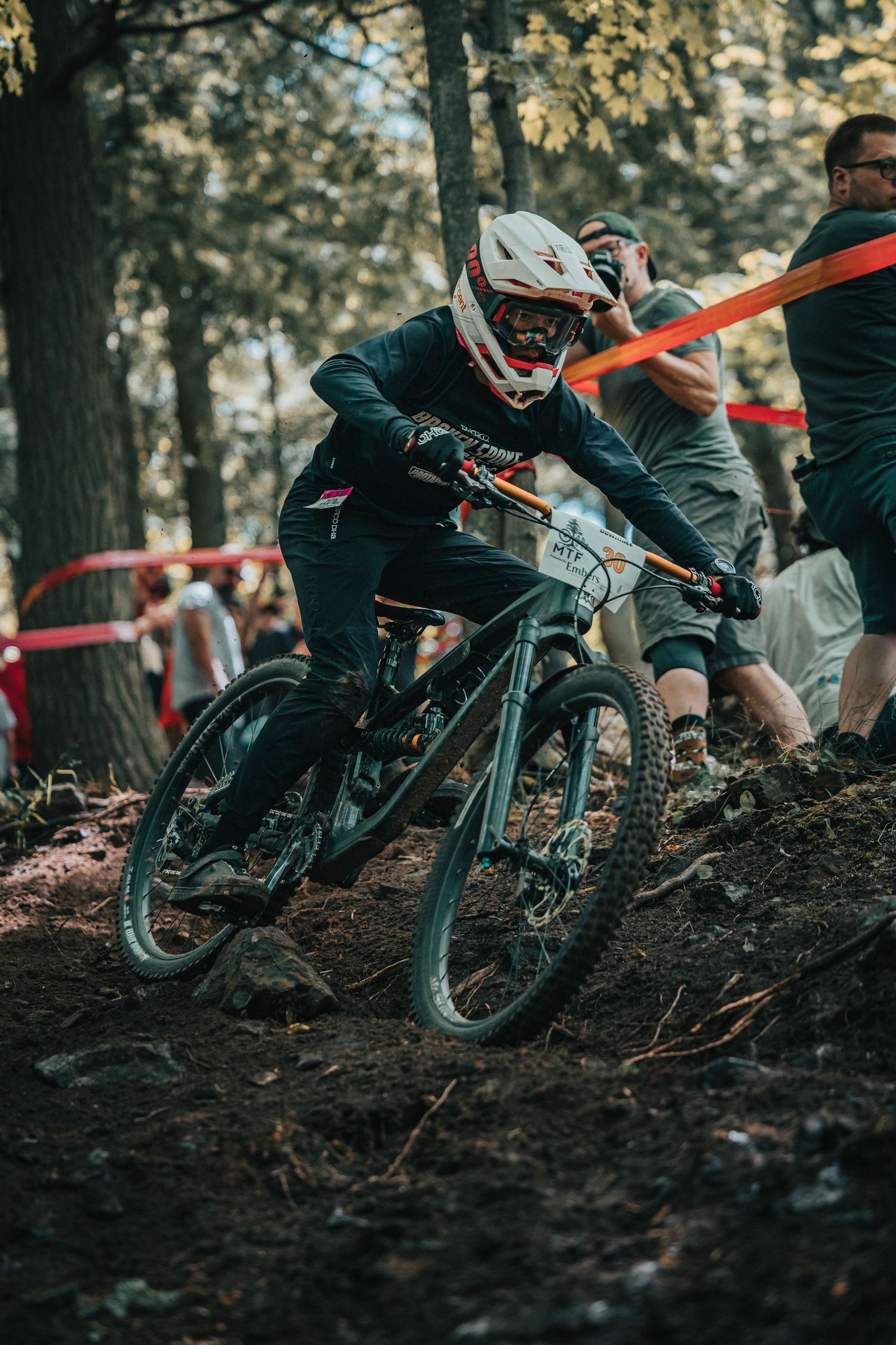 Mountain biker in white helmet and black gear races through a dirt forest trail past spectators.