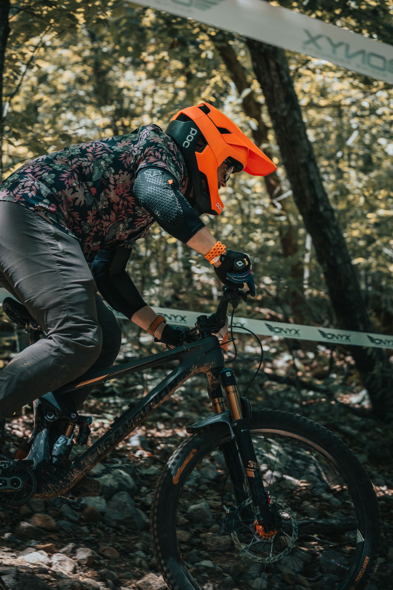 Mountain biker in orange helmet navigating rocky forest trail during downhill race.