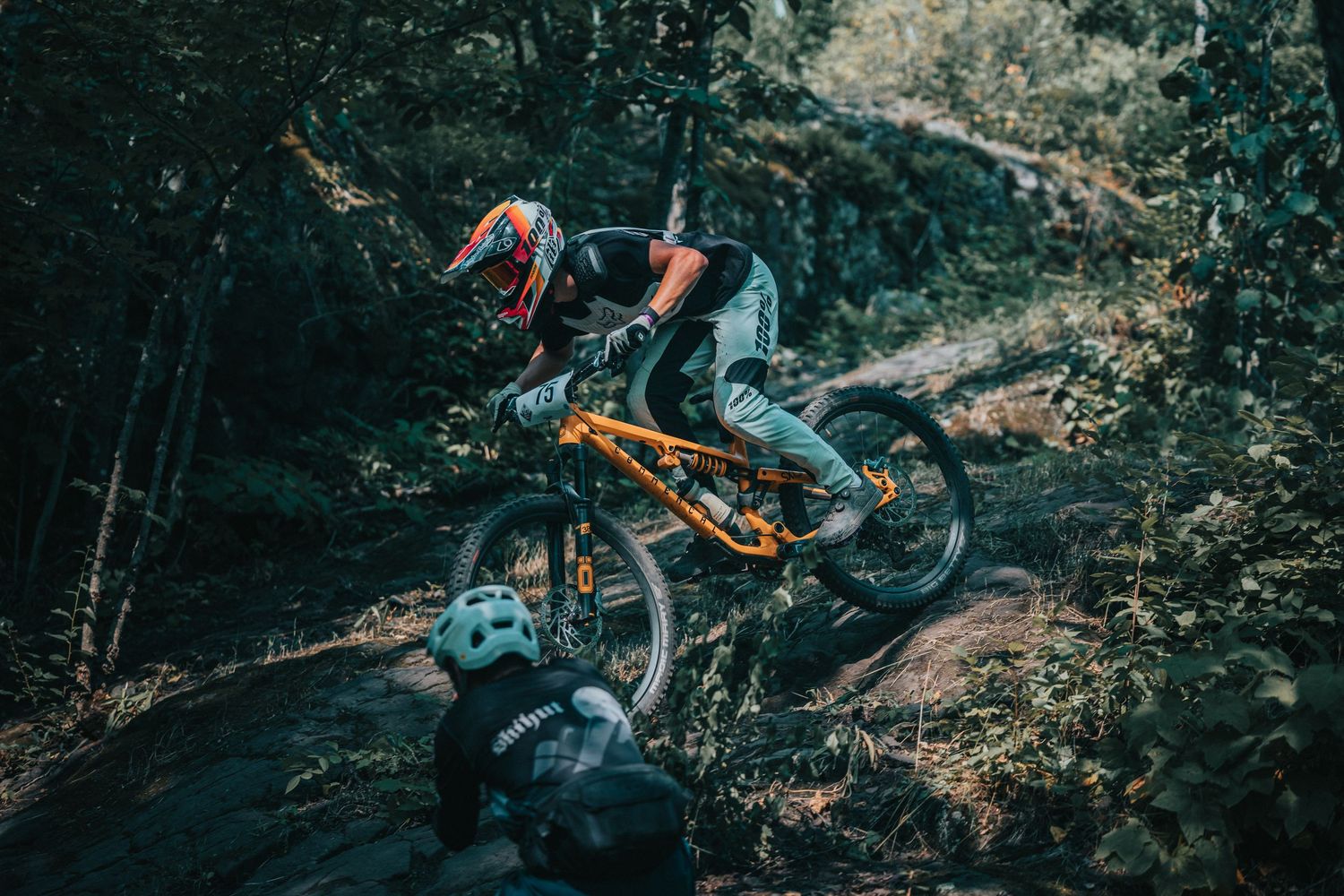 Two mountain bikers performing technical maneuver on steep rocky forest trail with orange bike.