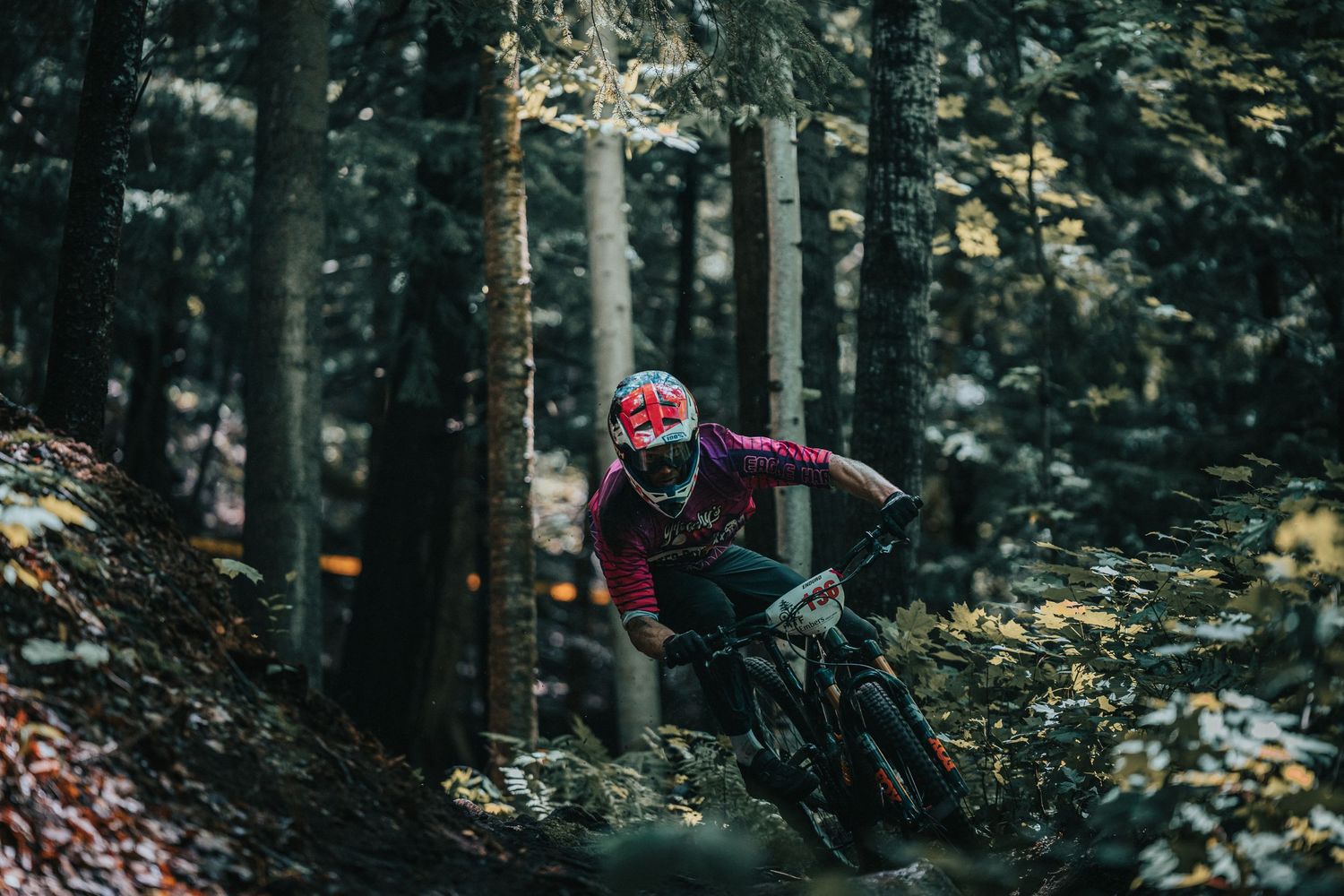 Mountain biker in red helmet riding downhill through dense forest trail at speed.