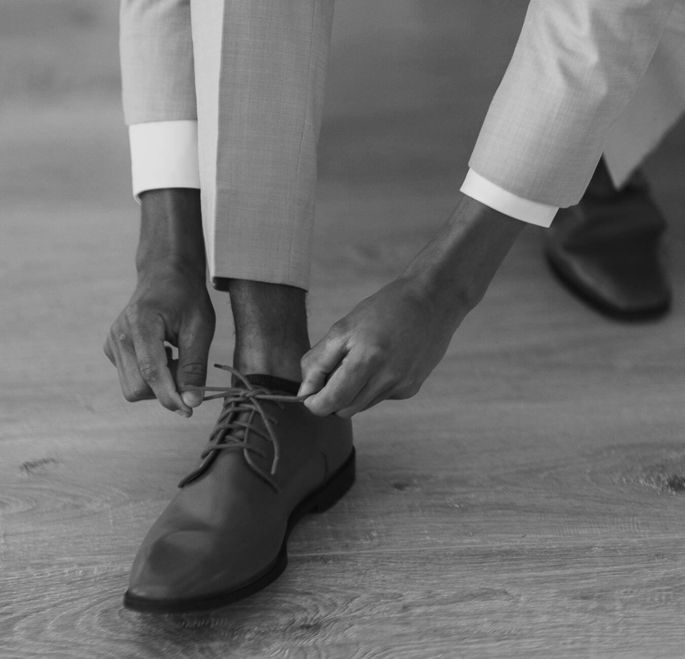 Black and white photograph of a groom leaning down to tie his dress shoes while wearing a wedding suit.