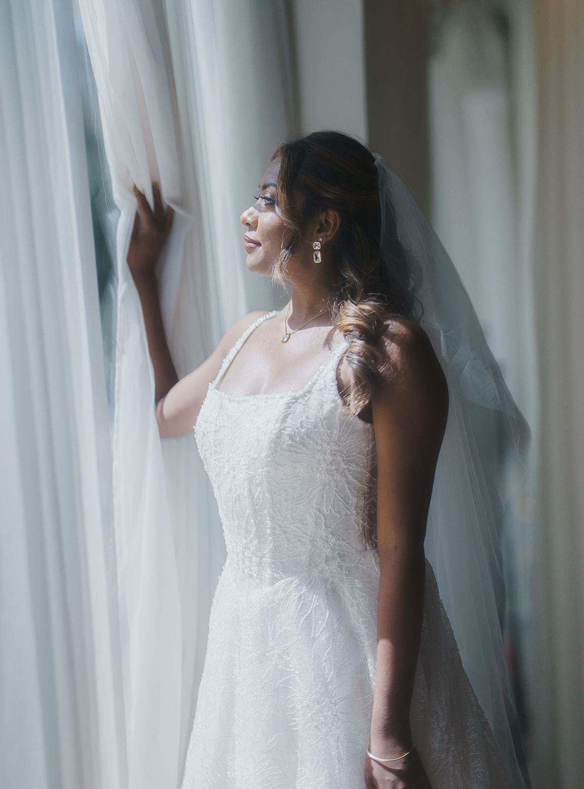 Bride in a white gown and veil looking out a sunlit window.
