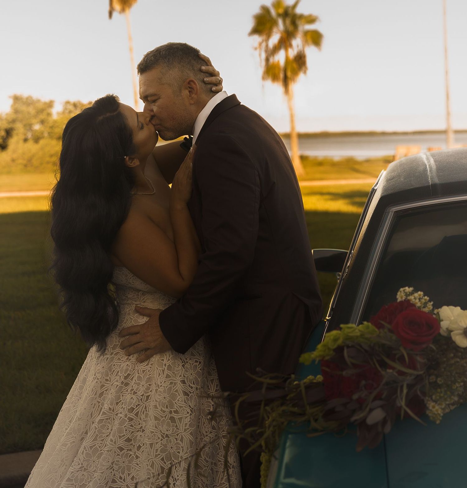 Bride in a lace dress and groom kissing while leaning against a classic vintage car decorated with red and white flowers under palm trees.