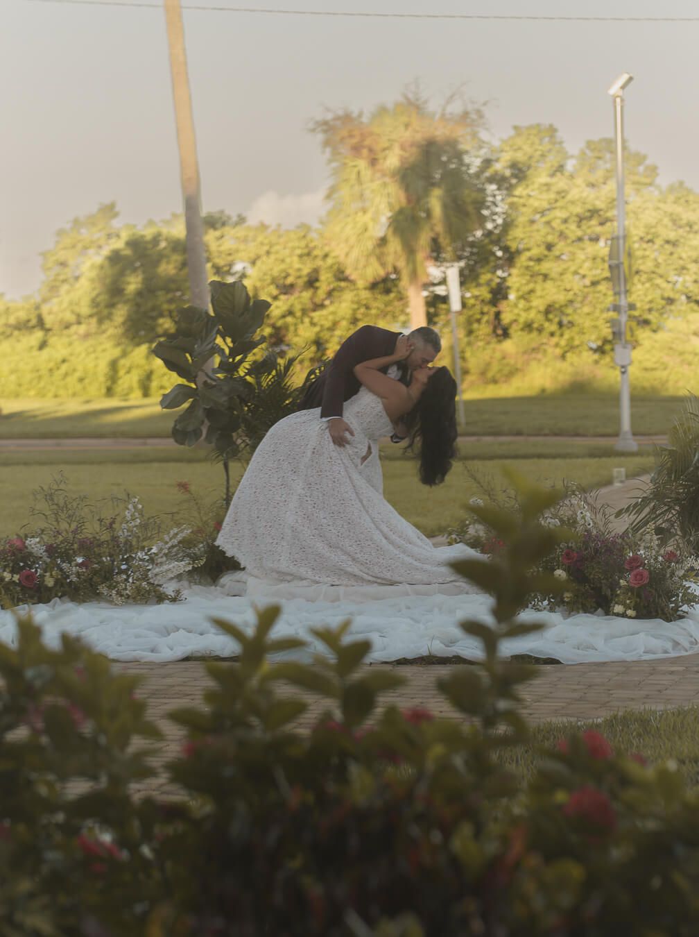 Bride and groom kissing in a dramatic dip surrounded by outdoor floral decor at sunset.