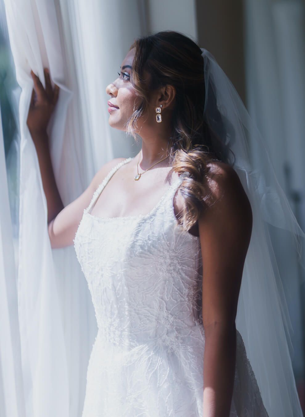 Profile shot of a bride in her wedding gown and veil holding back a sheer white curtain and looking out a bright window