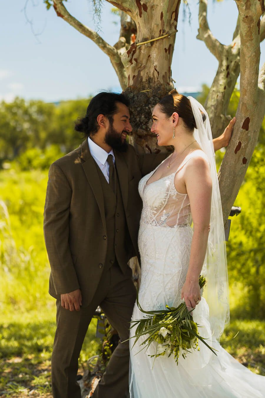 A bride holding a green and white bouquet smiles at her groom as they lean against a textured tree trunk in natural light