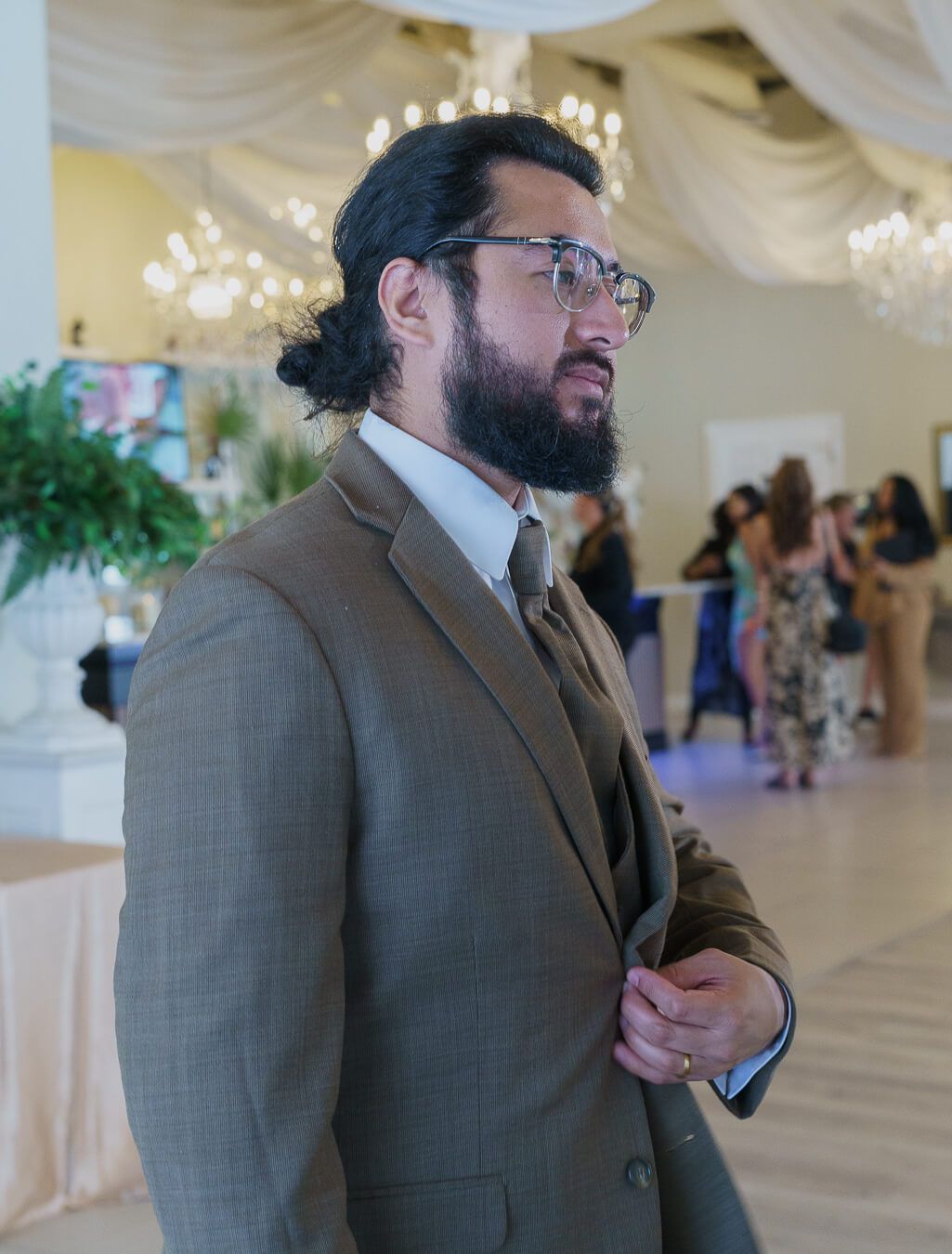 Side profile portrait of a bearded groom wearing glasses and a brown suit inside a wedding venue reception hall with chandeliers blurred in