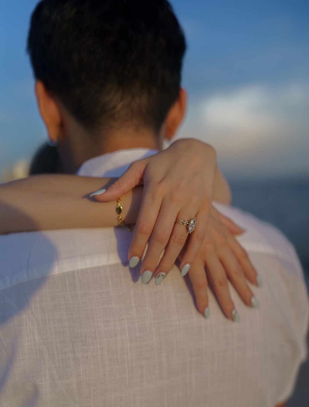 Close-up of a woman's hand wrapped around her partner's shoulders, showcasing a marquise cut diamond engagement ring against a soft blue sky