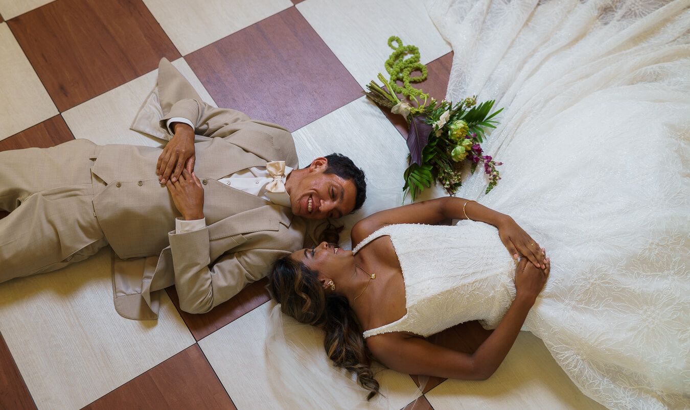 High-angle, overhead shot of a bride and groom lying down on a checkered floor next to each other, smiling