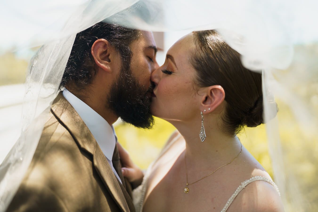 Close-up profile shot of a bride and groom kissing outdoors with her sheer white wedding veil draped over both of them