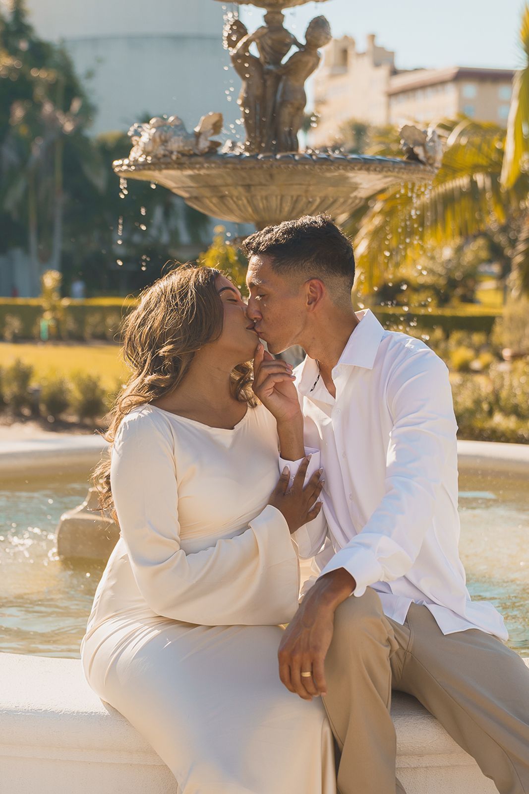 Man and woman kissing while seated by an outdoor water fountain in warm sunlight