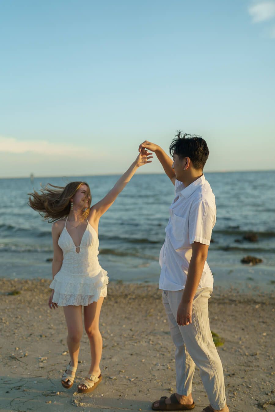A young couple dressed in white dancing and twirling on the shoreline at sunset