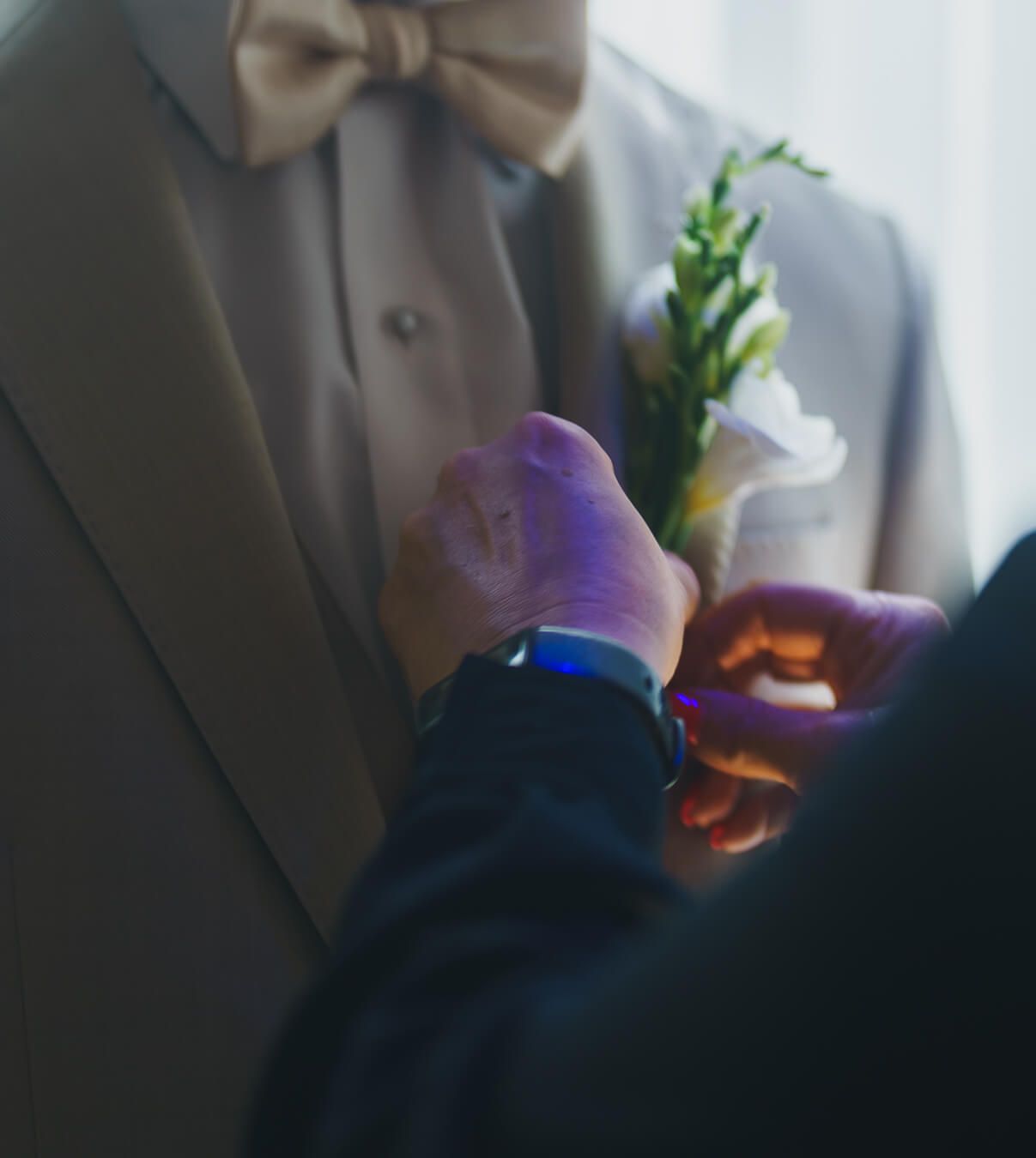 Close-up of a groom in a beige suit and bow tie having a white flower boutonniere pinned to his lapel.