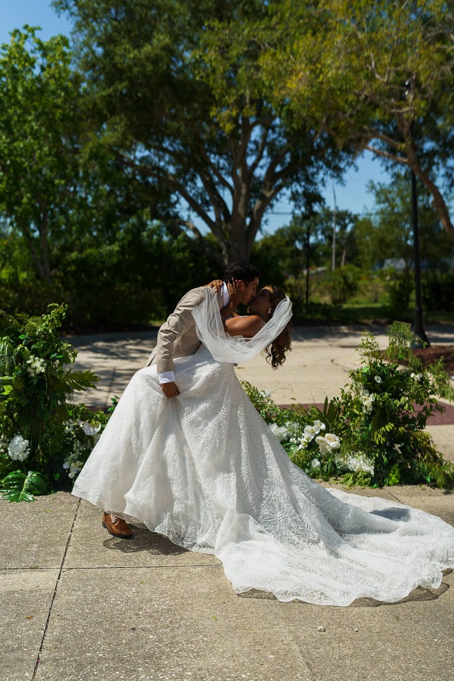 A groom dipping and kissing his bride outdoors with trees in the background, showcasing her flowing white wedding gown and veil.