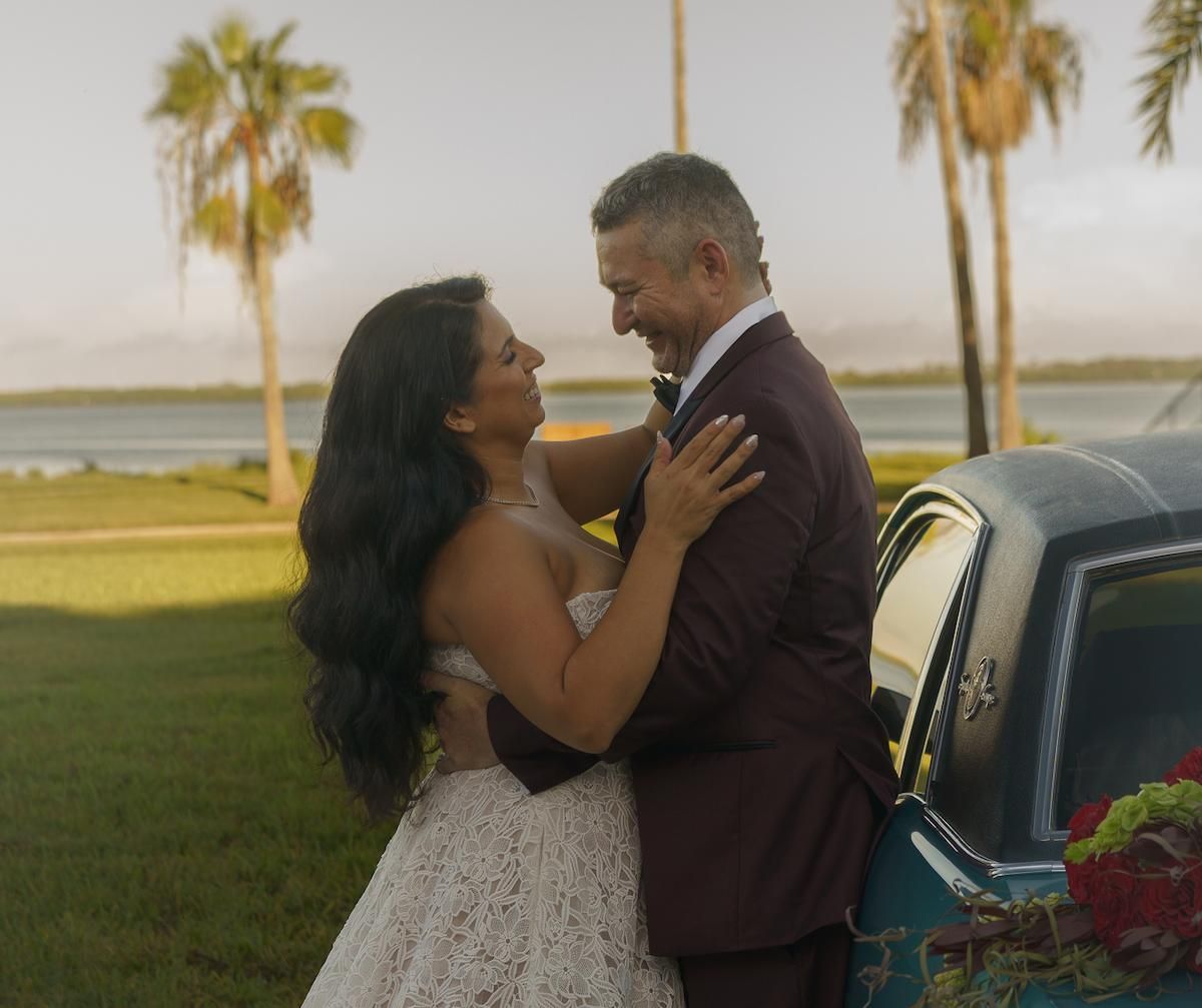Bride and groom sharing a laugh while leaning on a vintage car next to a palm tree shoreline.