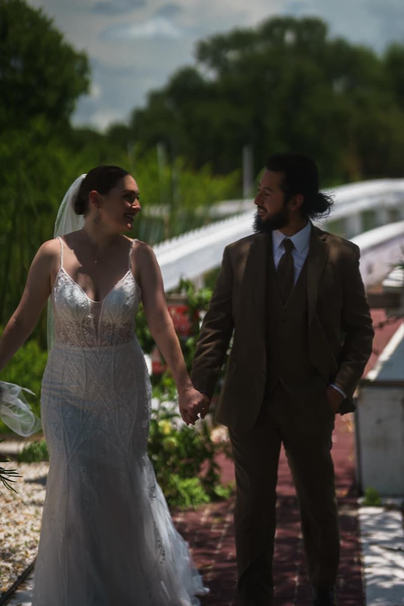 A smiling bride and groom holding hands and walking together along an outdoor brick pathway on a sunny day
