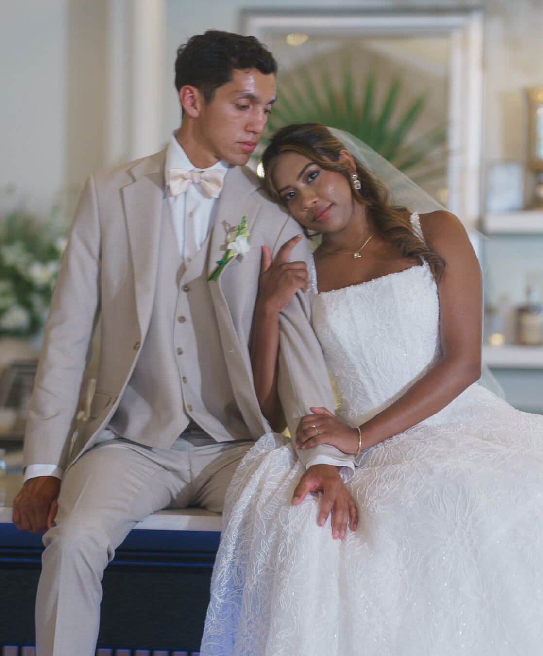 A bride in an intricate lace gown leans her head on her groom's shoulder while holding his hand, sitting together in a beautifully lit indoo