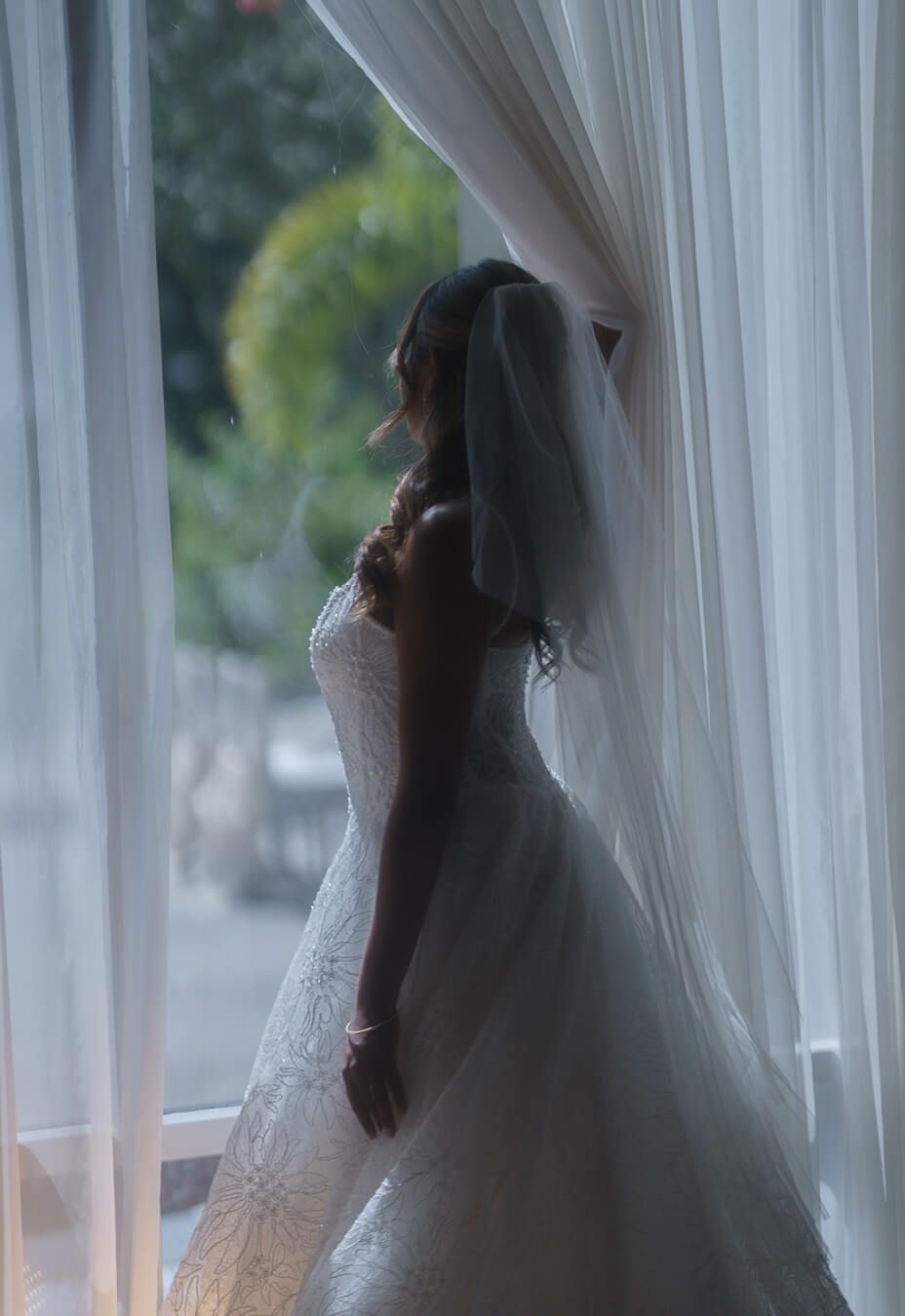 Artistic silhouette of a bride wearing a ballgown wedding dress and veil, looking out a large window into a garden