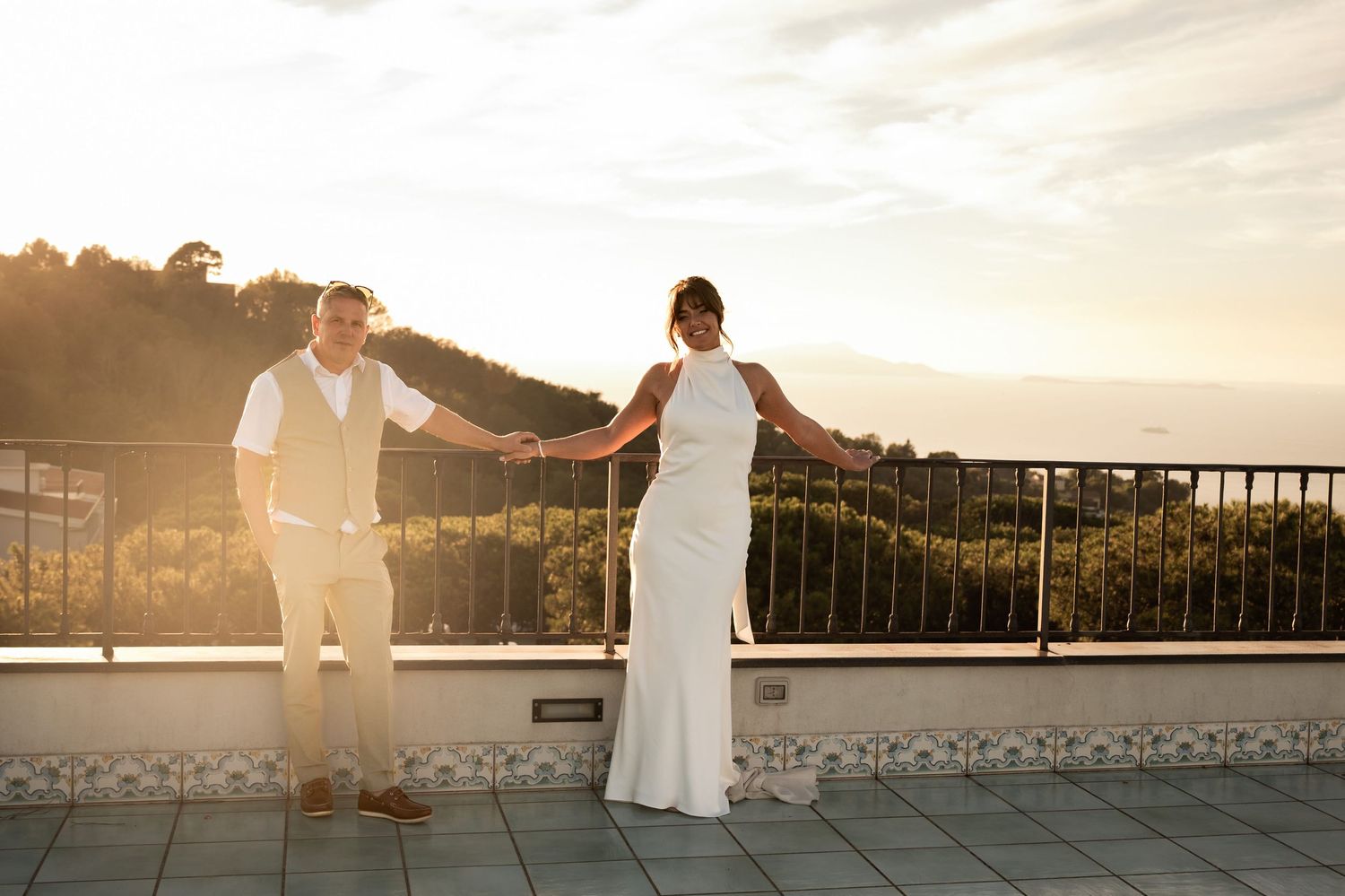A man and woman lean on the railings of a balcony holding hands, behind them a golden sun sets and creates a hazy orange glow.