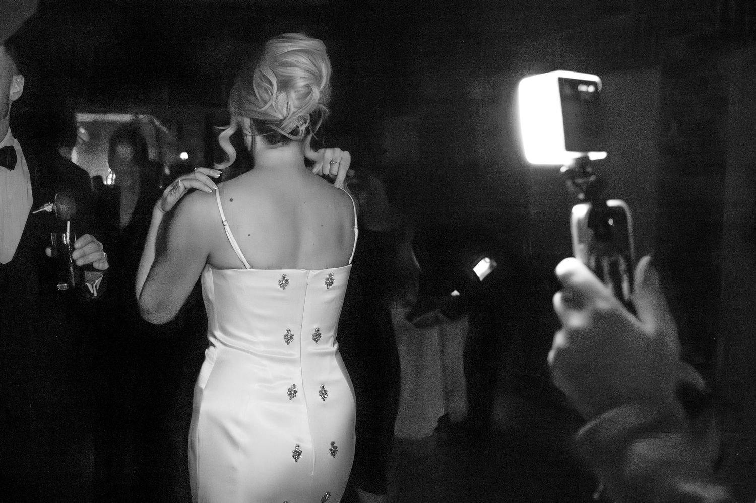 Woman photographed from behind with her hands rested on her shoulders. A bright handheld light illuminates her white jewelled dress.