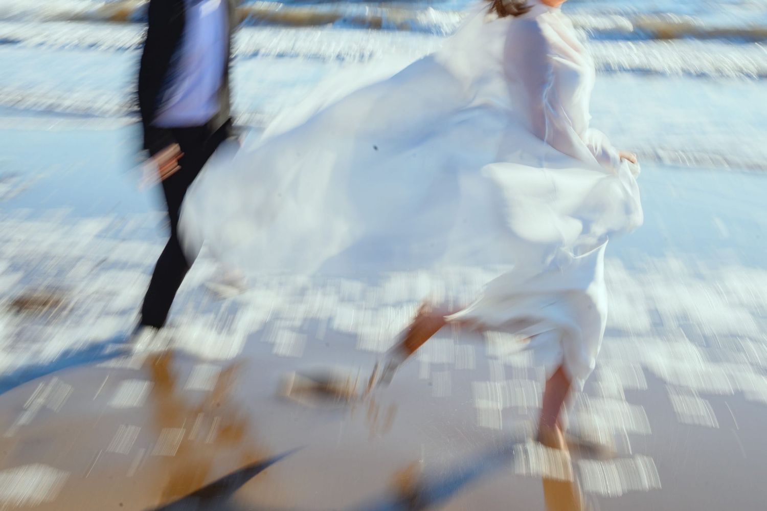 Woman holding up her white dress as she runs along the shoreline, whilst a man in a suit runs behind her.