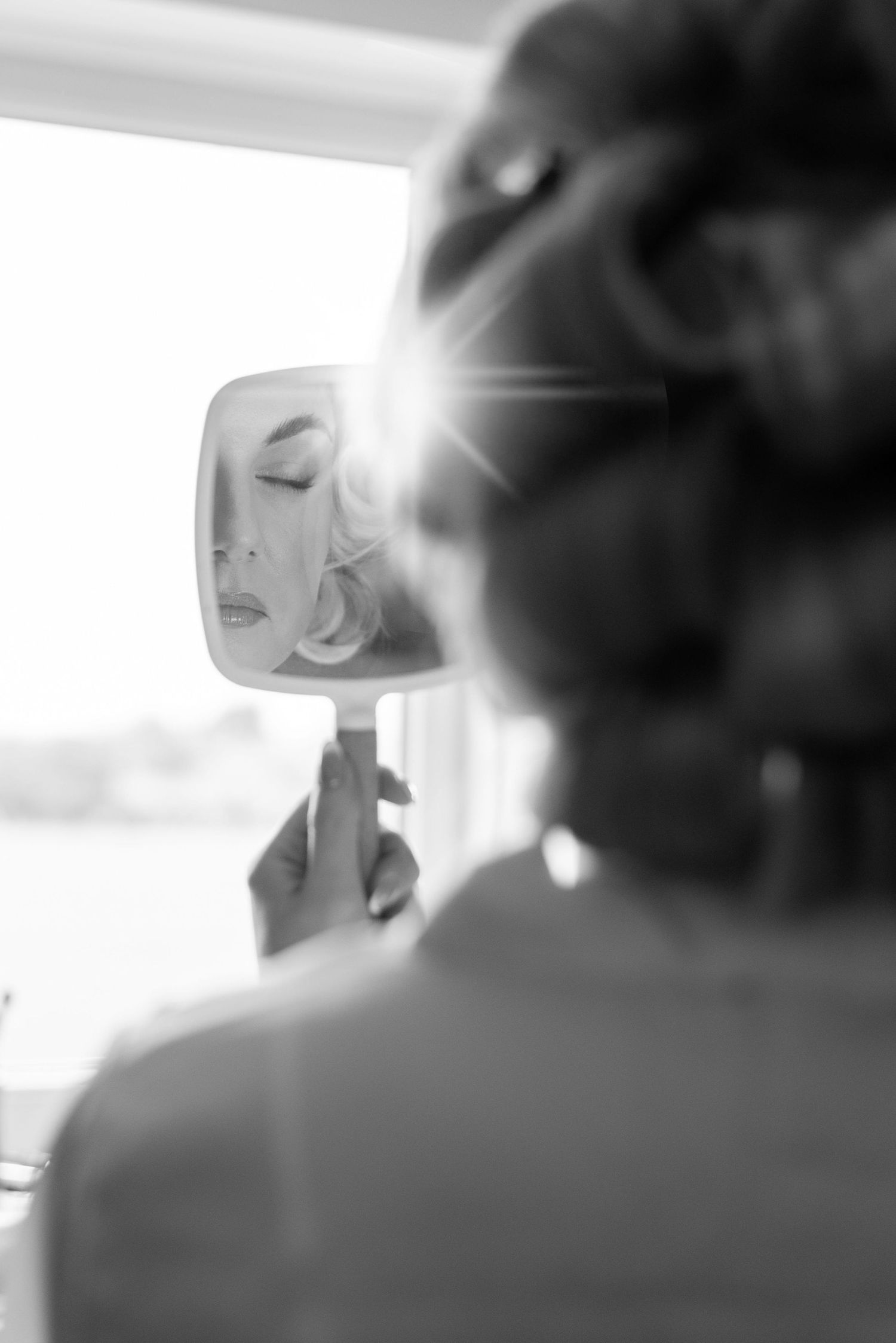 A woman standing in the window holding a small handheld mirror to her face. She has her eyes closed and rollers in her hair.