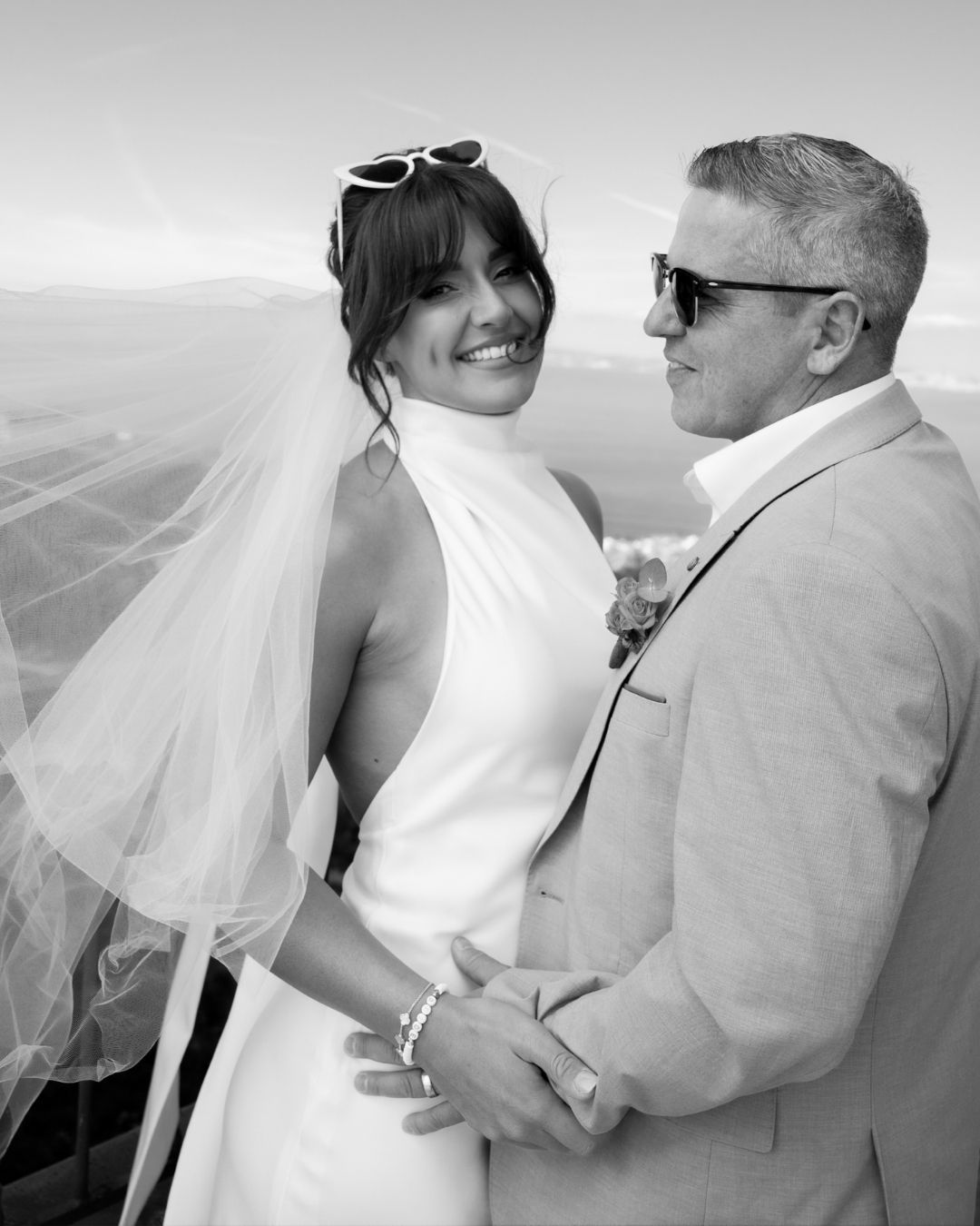 Happy brunette woman with white heart sunglasses on her head is held at the waist by a man wearing a suit with flowers on his lapel.
