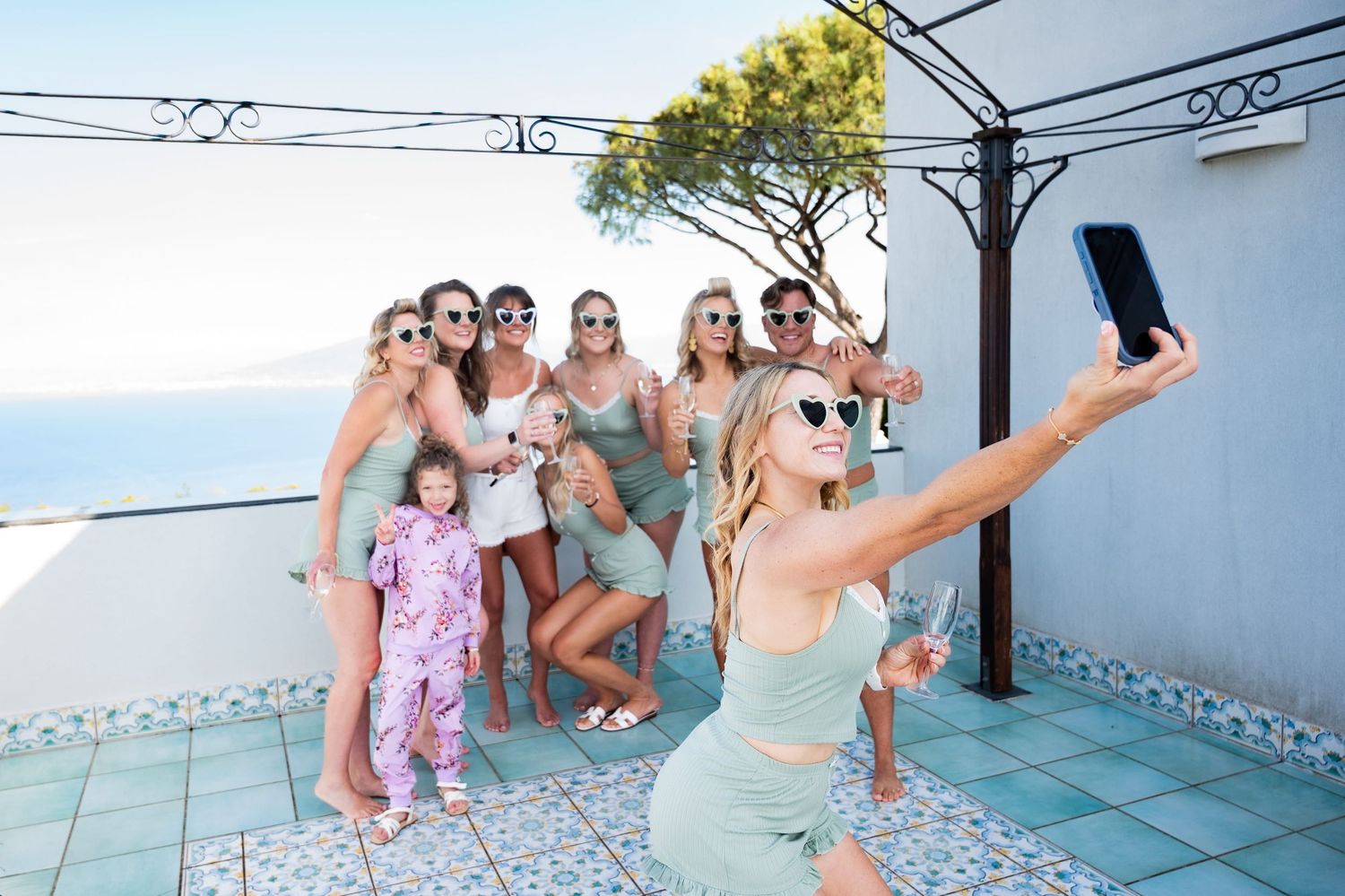 Wedding party posing together in matching sage green pajamas and heart shaped sunglasses. They are smiling and taking a selfie on a phone.