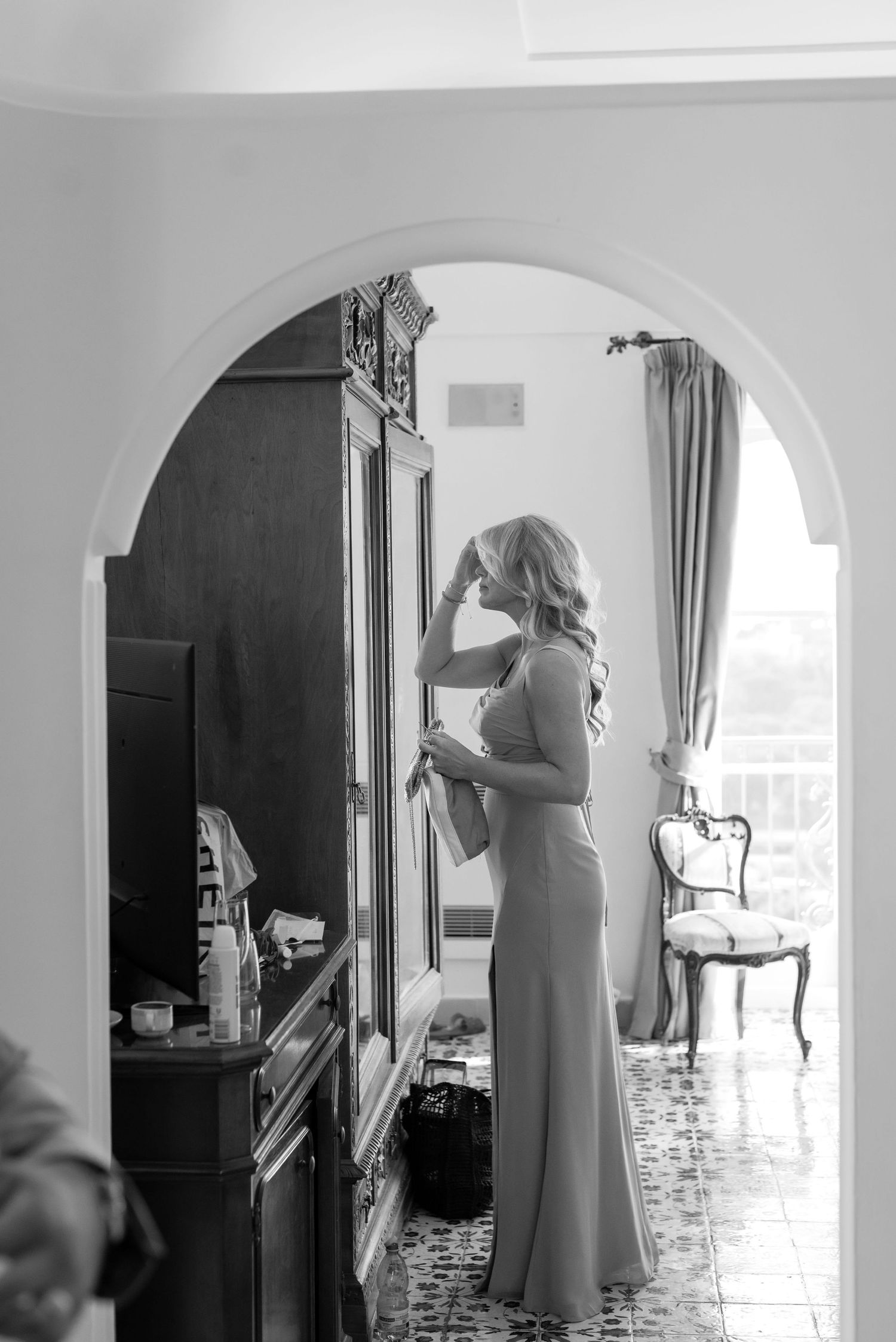 A bridesmaid standing in a room through an arch way. She is wearing a long gown and is looking in the mirror fixing her hair.