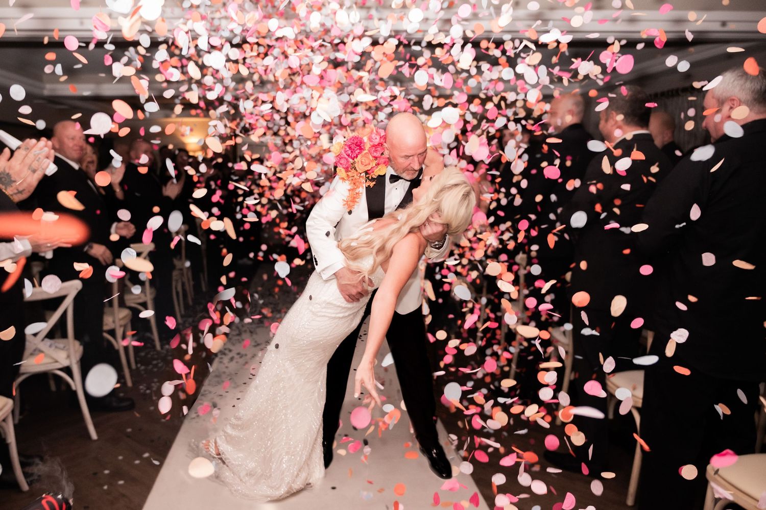A explosion of pink and white confetti engulfs a newly married couple, the groom leans down to kiss his bride as the congregation applaud.