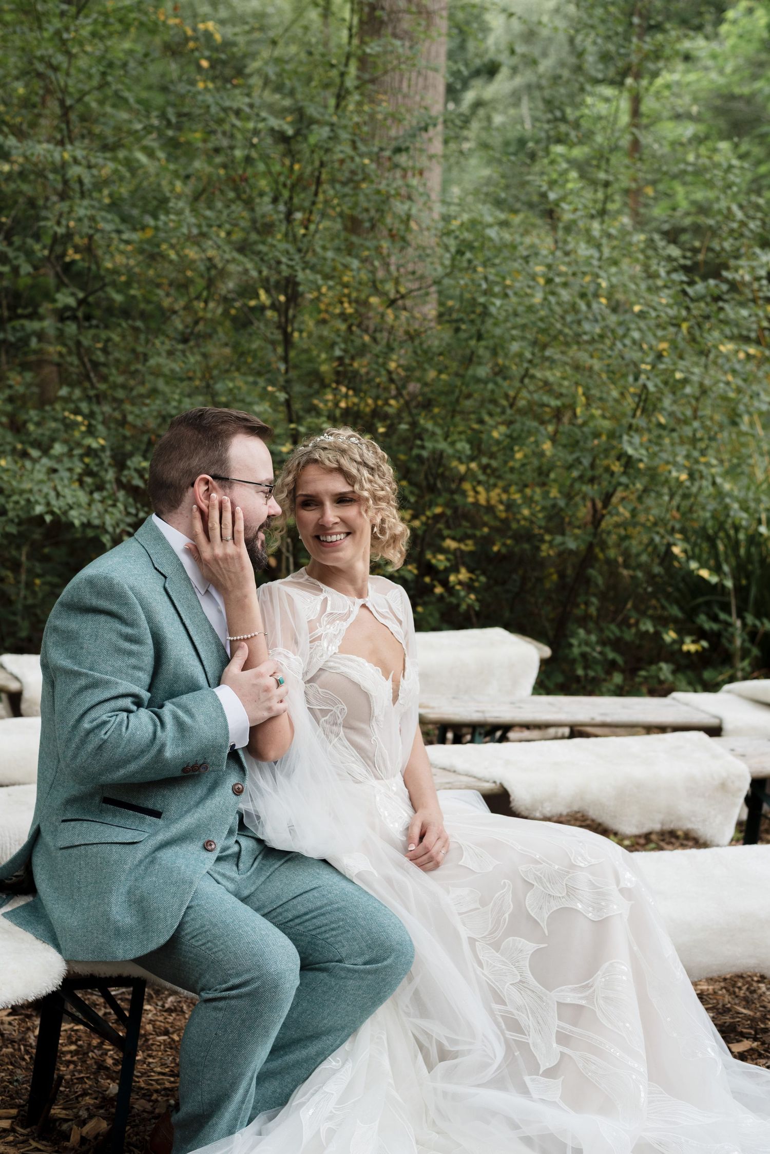 Couple sitting on blanketed benches in the forest, the man rests his hand on the woman's arm whilst she puts hers on his face.