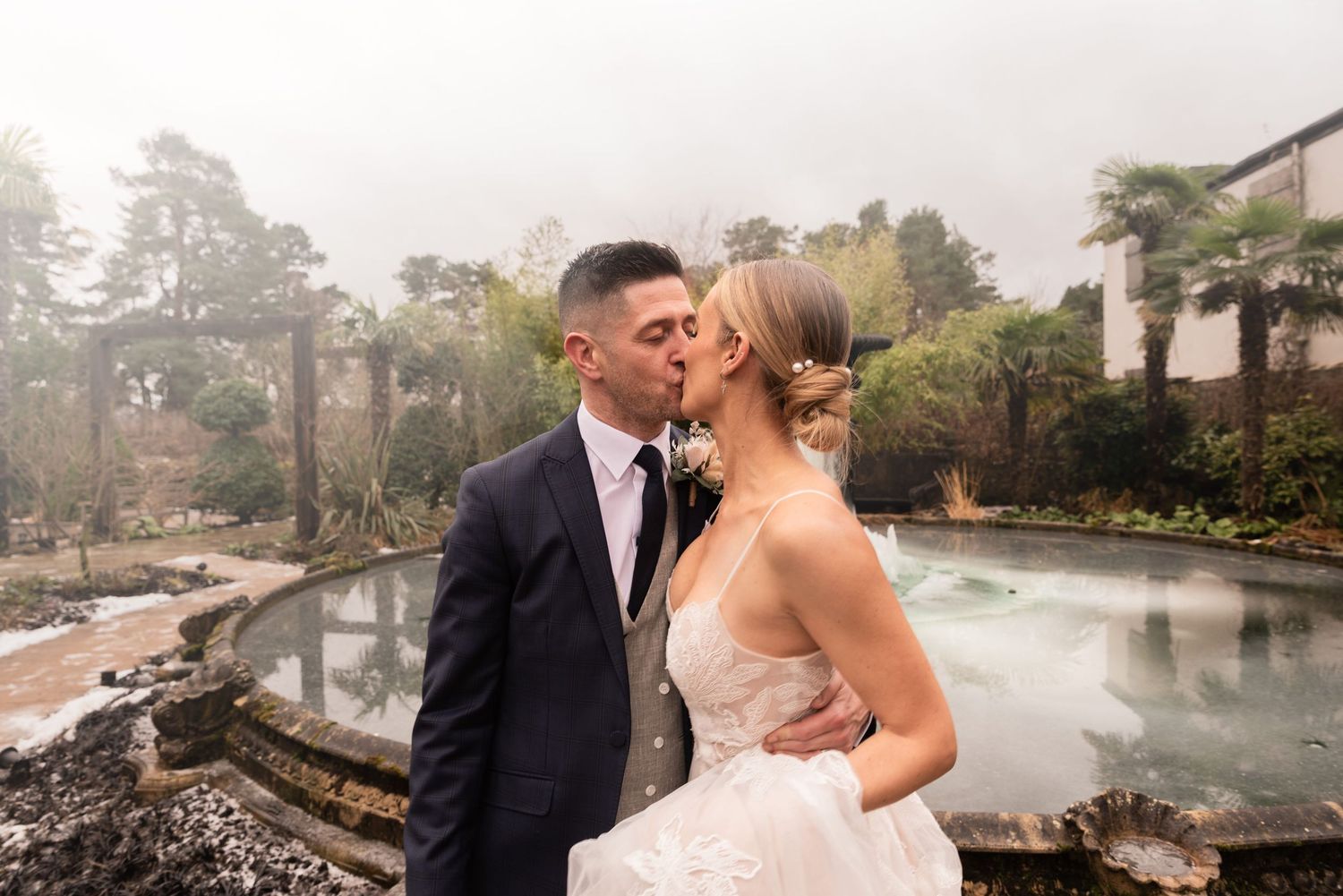 A couple embrace and kiss on a cold day in front of a frozen pond. Snow lies on the ground and mist engulfs the air and trees around them.