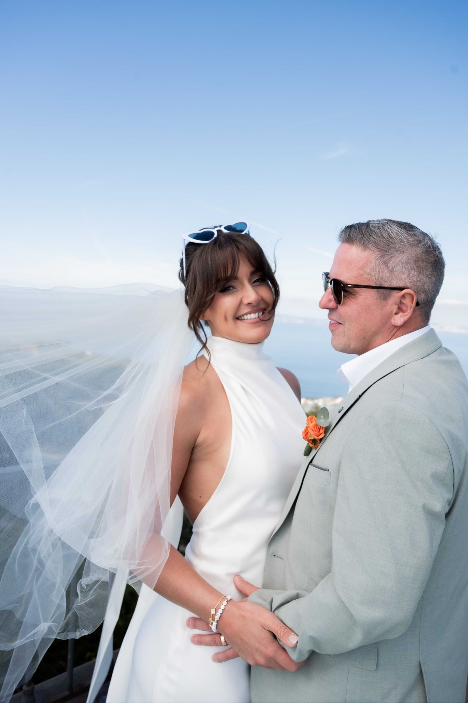Beaming bride holds hands with her husband on the coast, they stand close together and the wind blows her veil forward from behind her.