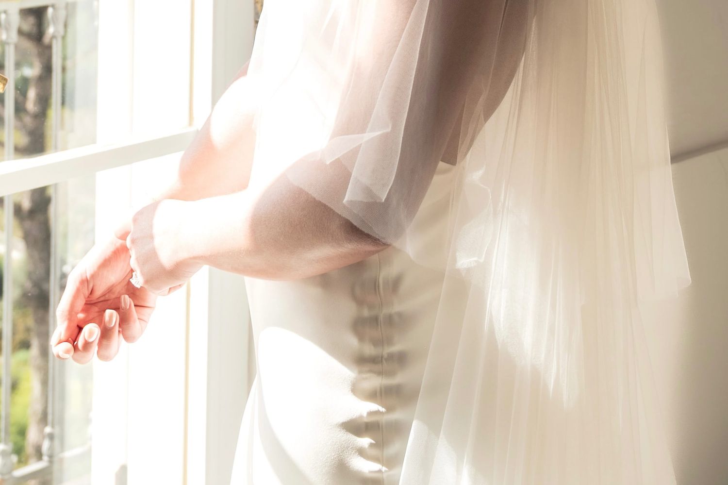 Detail shot as a bride adjusts jewellery on her wrist. She stands in front of a glass door and the sun shines in on her dress and veil.