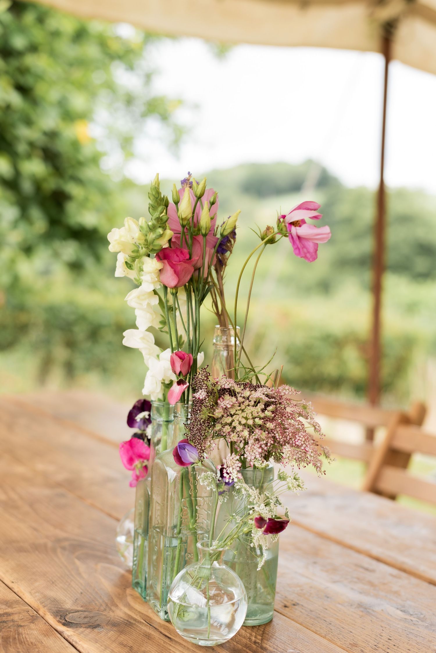 An arrangement of purple and white wildflowers in various different height glass bottles on a wooden table in a countryside setting.