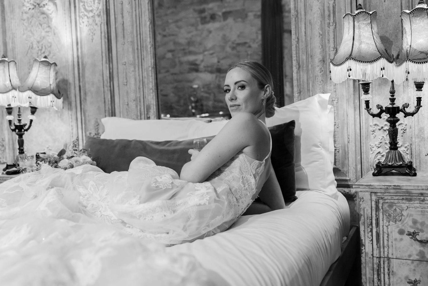 Woman sitting on a bed in an elaborately decorated hotel room holding a glass of champagne and wearing a long lace dress.
